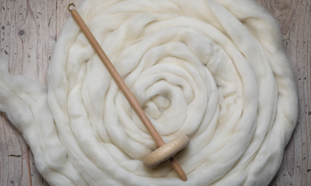 Weiße Schafwolle liegt zu einer Spirale aufgerollt flach auf einem hellbraunen Holz-Hintergrund. Auf der Wolle liegt eine hölzerne Spindel.